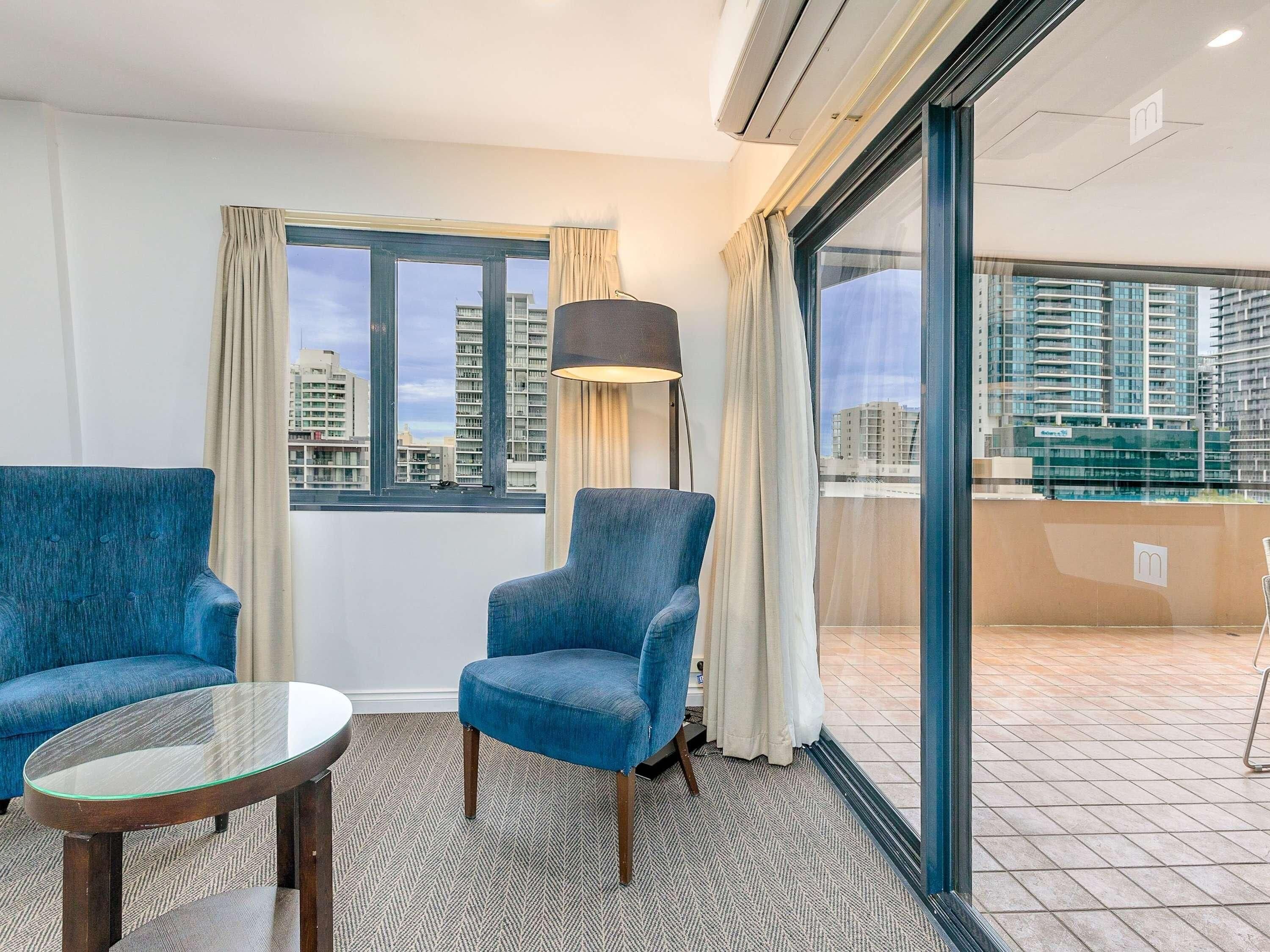 Habitación de hotel con dos sillones azules, una mesa de vidrio, una lámpara de pie y vistas a edificios altos a través de amplios ventanales.