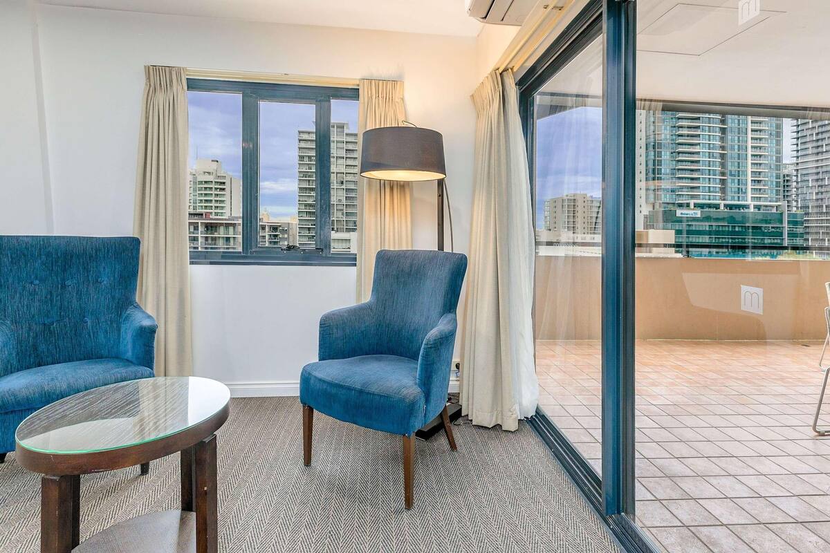 A hotel room with two blue armchairs, a glass table, a floor lamp, and a view of high-rise buildings through large windows.