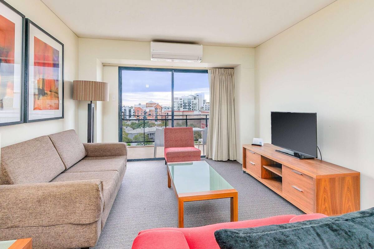 A hotel room with a sofa, armchair, TV, and a balcony with a city view.