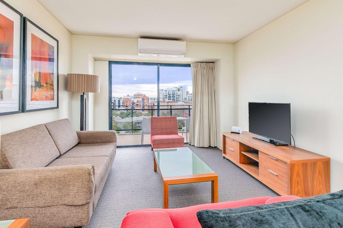 Habitación de hotel con sofá, sillón, televisión y balcón con vistas a la ciudad.