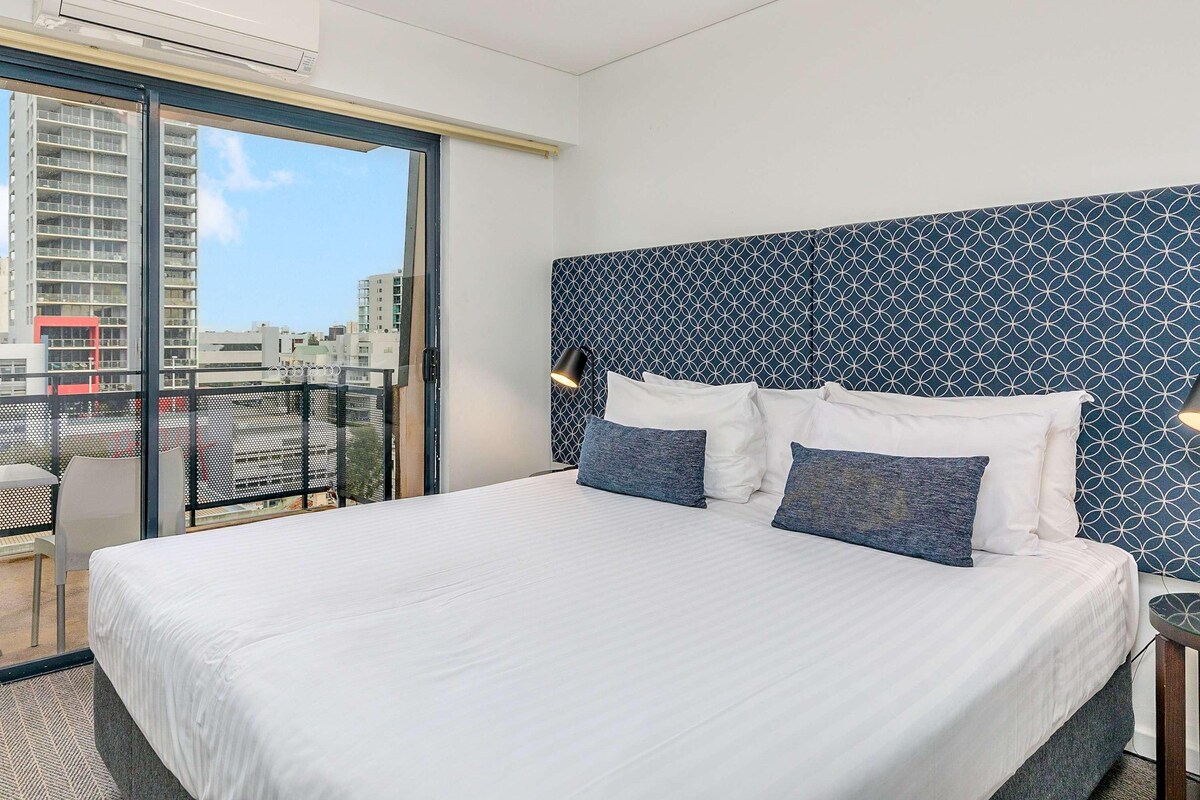 Une chambre d’hôtel avec un grand lit, une tête de lit décorée, un balcon offrant une vue sur la ville et un appareil de climatisation.