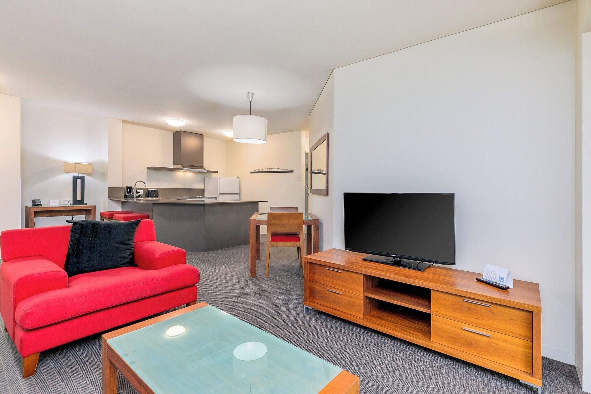 A modern hotel room with a red sofa, a glass coffee table, a TV on a wooden stand, a kitchenette, and a small dining area.