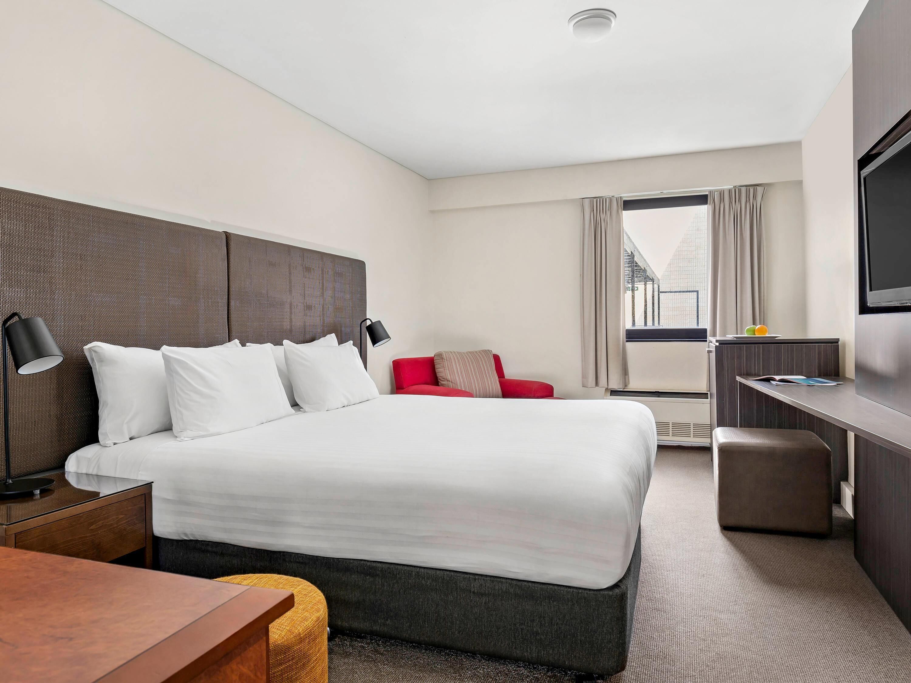Habitación de hotel con una cama grande, un escritorio, una silla roja, un televisor y un ventanal con cortinas.
