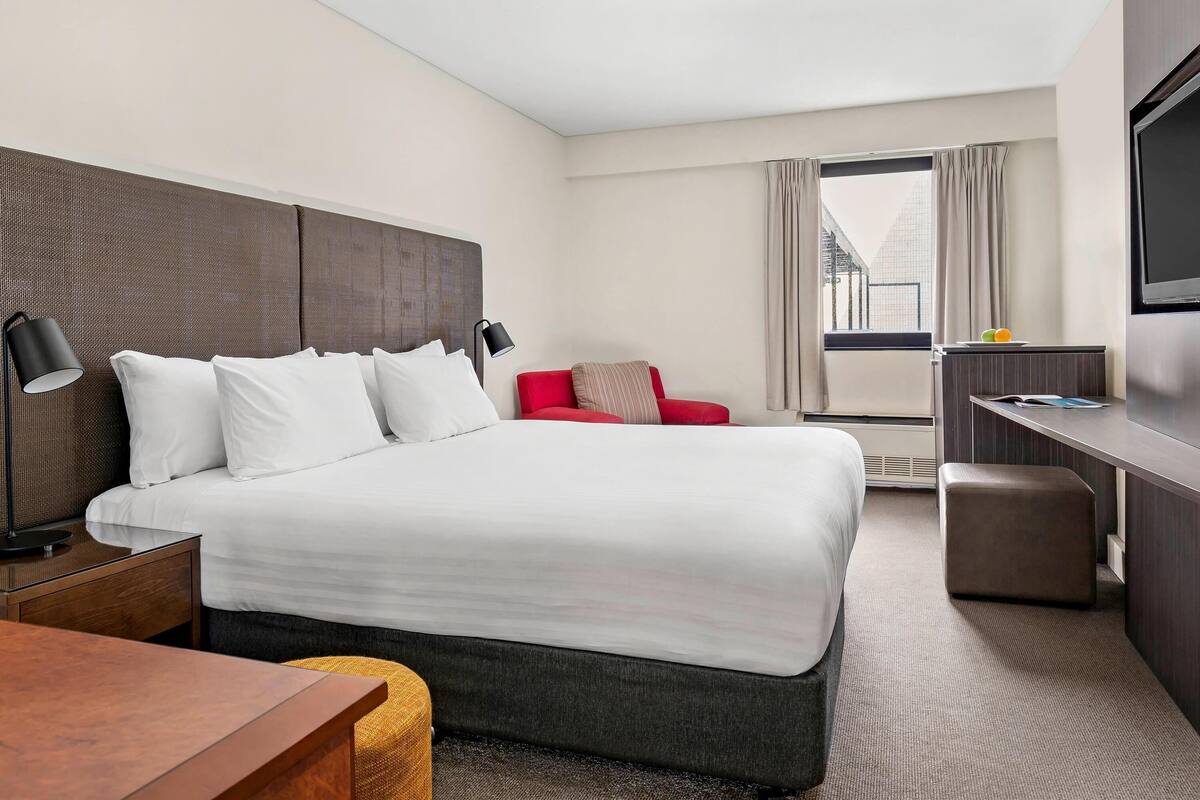 A hotel room with a large bed, a desk, a red chair, a television, and a window with curtains.