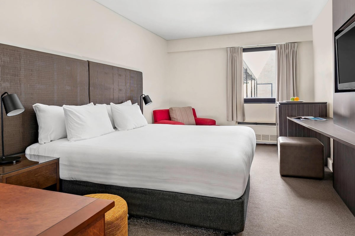 Habitación de hotel con una cama grande, un escritorio, una silla roja, un televisor y un ventanal con cortinas.