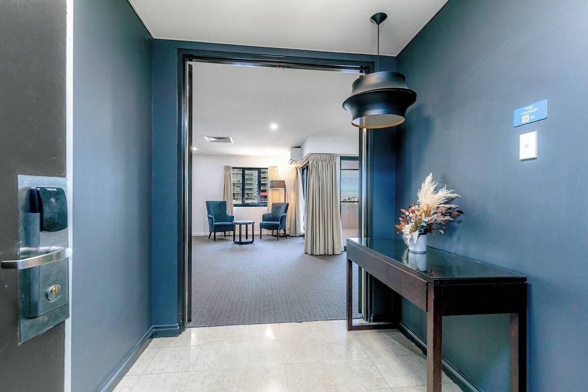 A hotel hallway with a dark blue wall, a modern light fixture, a small table with a vase of dried flowers, and a glimpse of a dining area in the background.