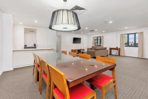A modern hotel room with a large dining table, wooden chairs, a sofa, a TV, and a large window with curtains.