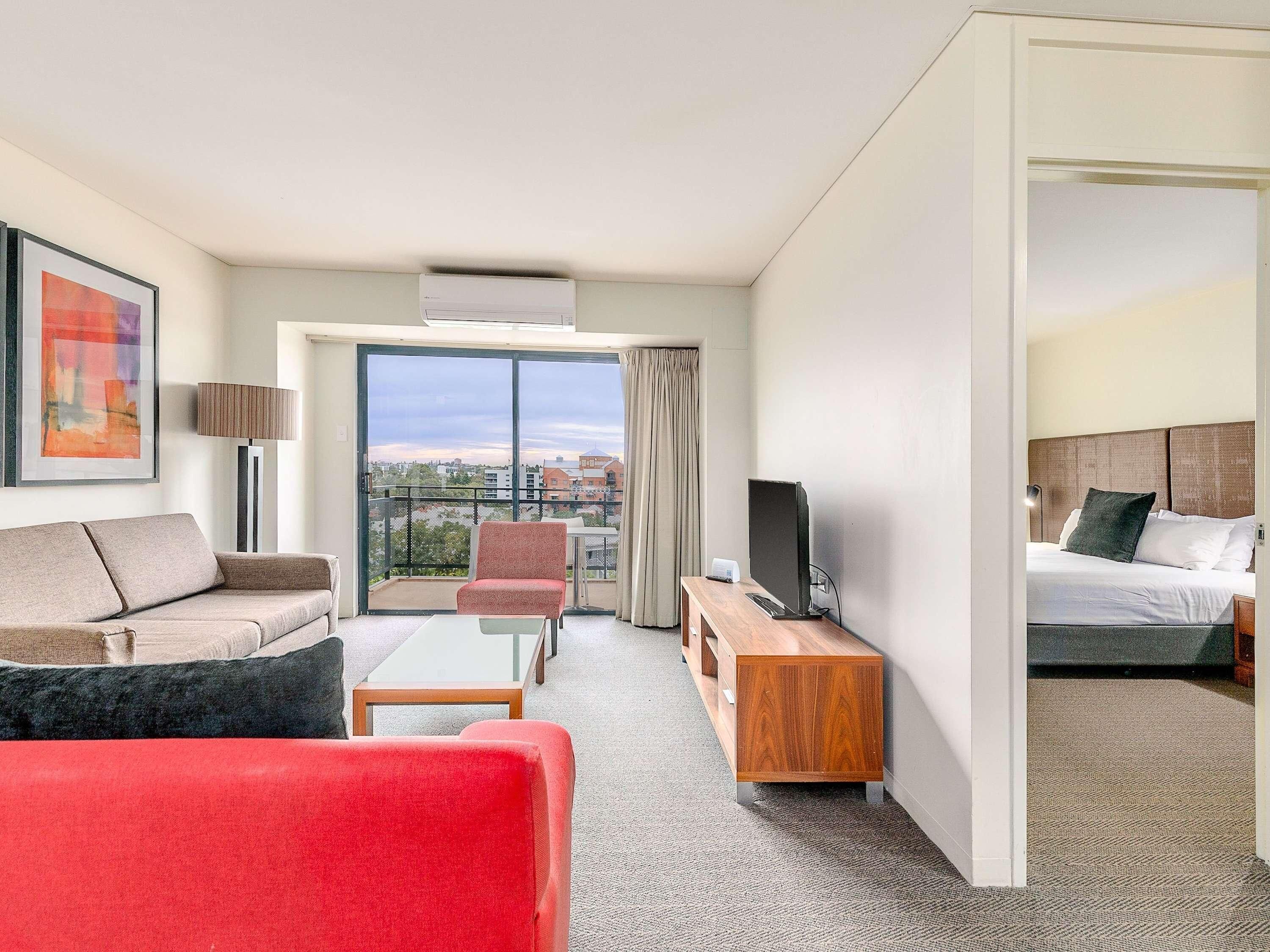 Habitación de hotel con sofá, una silla roja, televisor sobre un mueble de madera, cama y balcón con vistas a la ciudad.