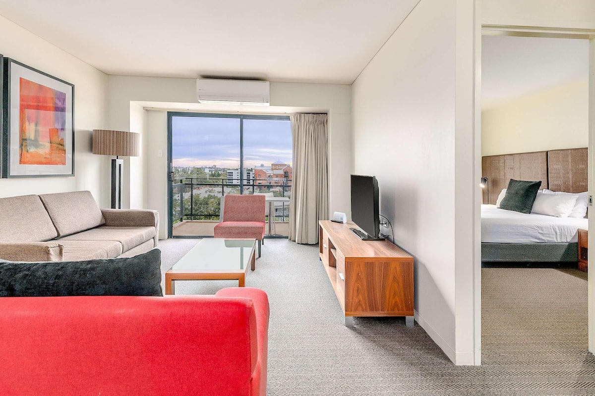 Habitación de hotel con sofá, una silla roja, televisor sobre un mueble de madera, cama y balcón con vistas a la ciudad.