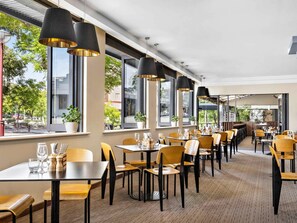 Un restaurante con mesas y sillas, ventanas amplias y lámparas colgantes.