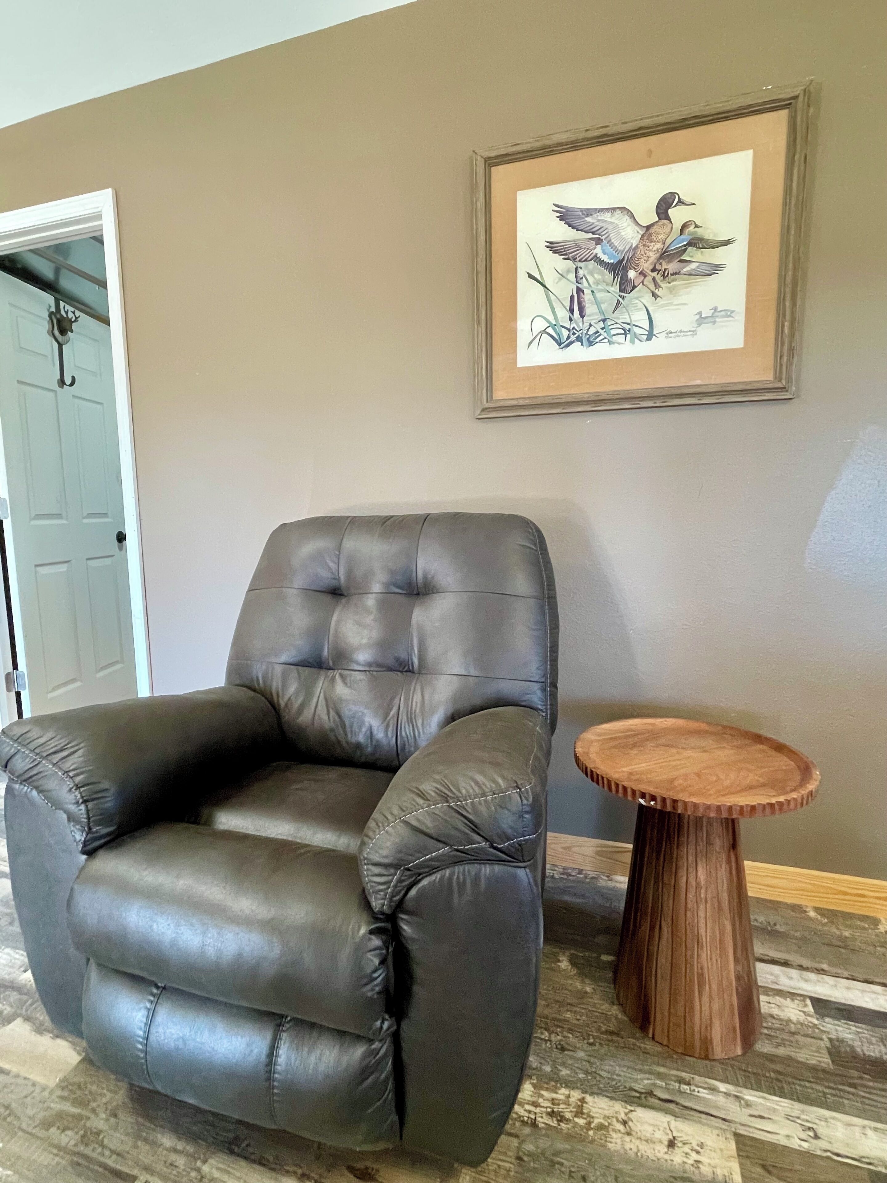 A dark leather armchair with a wooden side table in a room with a beige wall and a framed picture of birds.