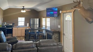 A modern living room with a fireplace, a mounted deer head, and a kitchen area with a TV. - Country Cabin in Private Setting (Altha)