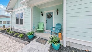 A light blue house with a porch, two green Adirondack chairs, and potted plants.