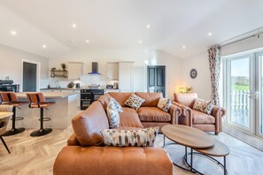 A modern living room with a sofa set, a coffee table, and a kitchen area with a stove and cabinets. - 3 Bed in Plympton (oc-m30351) (Sparkwell)