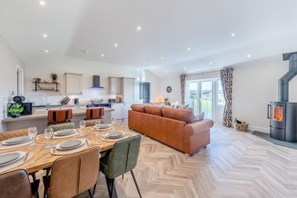 A modern kitchen with white cabinets, a dining area with a wooden table set for four, and a large sofa in the living room.