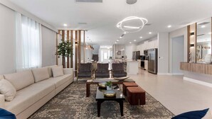 Uma sala de estar moderna com um sofá, cadeiras, uma mesa de centro e uma cozinha ao fundo.