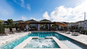 Uma piscina retangular com uma banheira de hidromassagem no centro, rodeada por espreguiçadeiras e guarda-sóis.