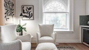 A living room with two armchairs, a side table, a potted plant, a framed botanical print, and a window with a decorative glass design. - Plums Unit 2: Downtown - King (Stratford)