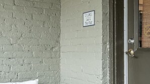 A corner with a white wicker chair, a brick wall, and a door with a sign reading "DLUMS UNIT 4 The Flat".