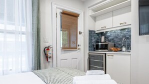 A compact hotel room with a bed, a kitchenette, and a door with a window blind.