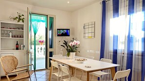 Un coin repas avec une table blanche, des chaises blanches, un fauteuil à bascule en osier, un téléviseur fixé au mur et une vue à travers une porte vitrée.