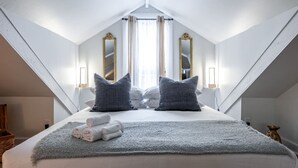 A bedroom with a high ceiling, a large bed with pillows, a bedside table, and two wall-mounted lamps. - Plums Unit 3: Downtown King (Stratford)
