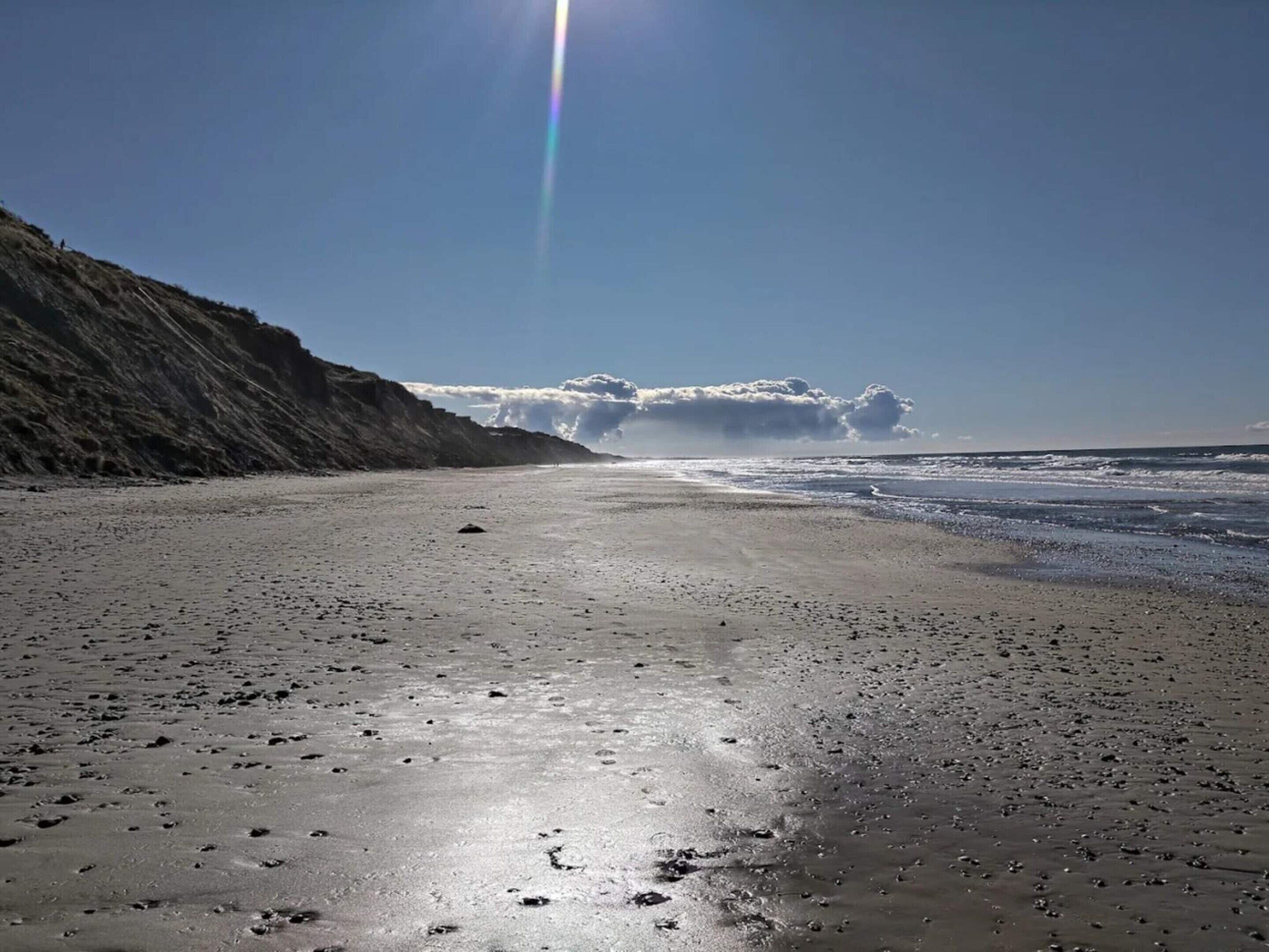 Een strand met een heldere lucht, een zichtbare regenboog en een klif aan de linkerkant.