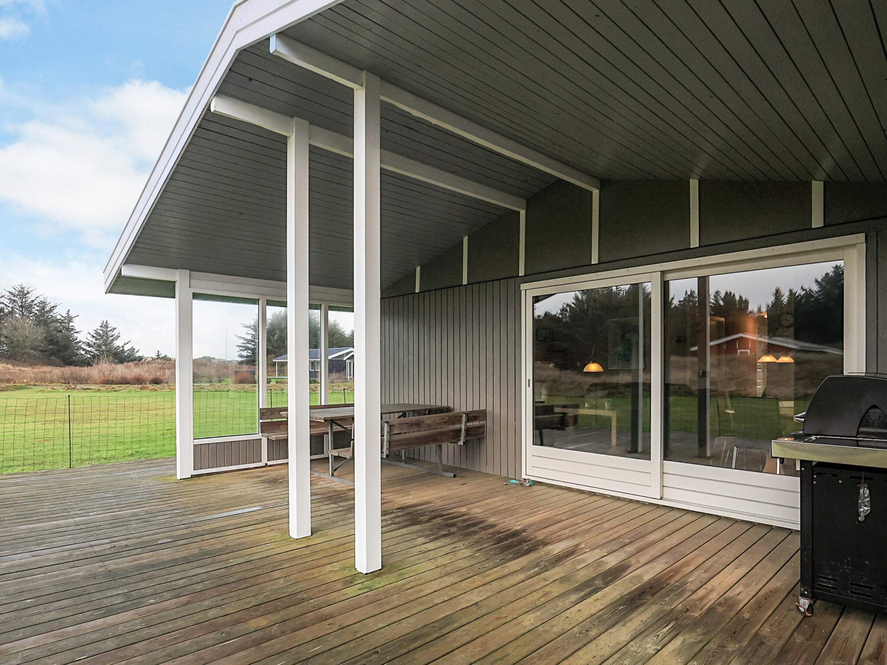 Een overdekt buitengebied met een houten terras, een barbecue en een glazen deur die uitkomt op een groene plek.