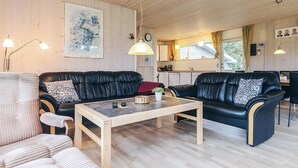 Una sala de estar acogedora con un sofá de cuero negro, una mesa de centro de madera y un sillón a rayas. Se ve una cocina al fondo, con una ventana y cortinas.