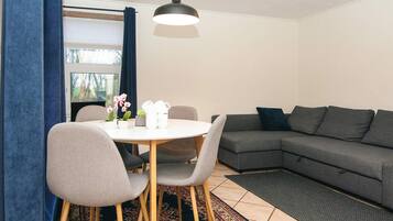 Una sala de estar moderna con un sofá gris, una mesa de comedor redonda con sillas, una lámpara colgante y una ventana con cortinas.