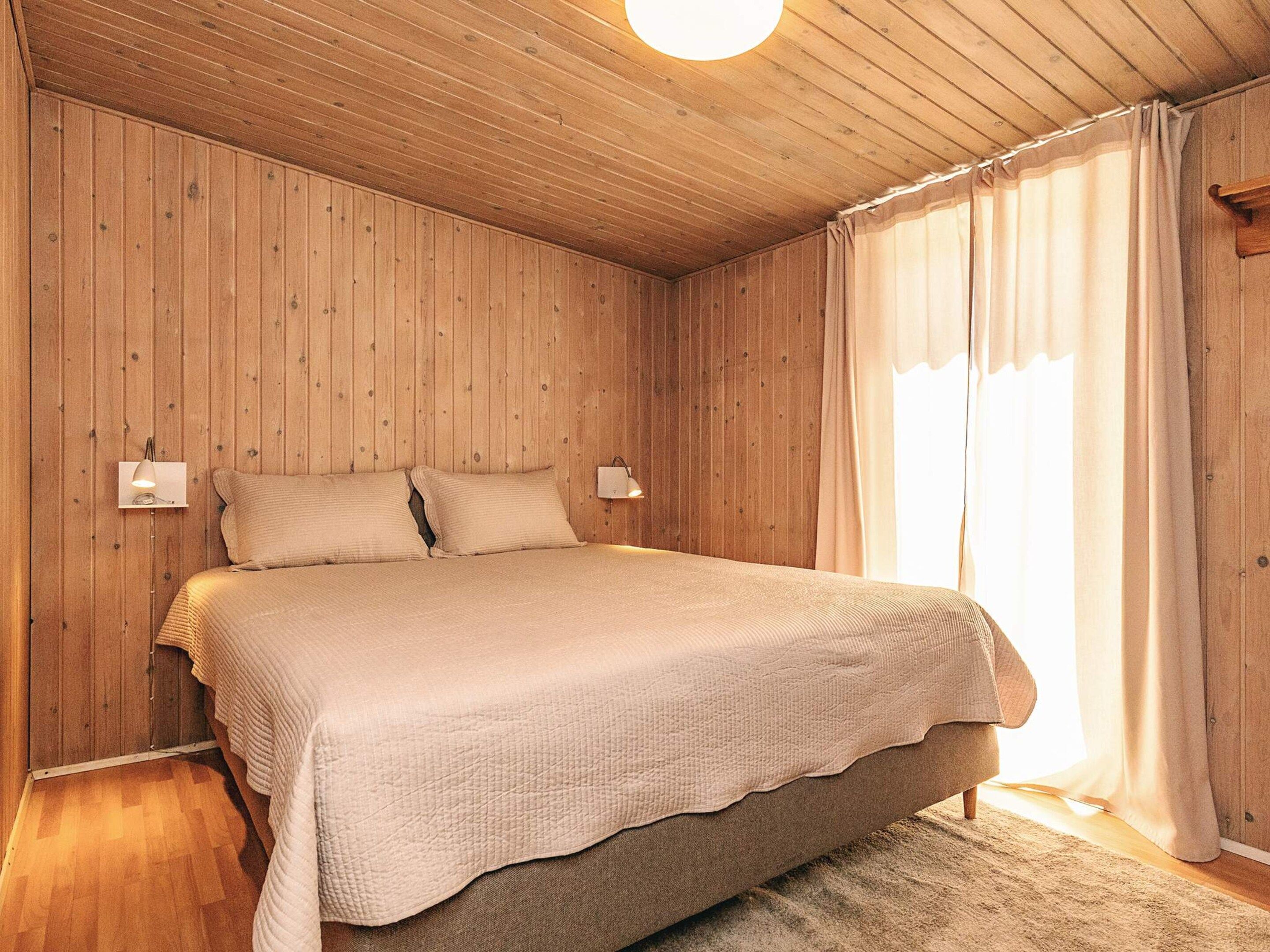 Een slaapkamer met houten paneelwanden, een bed met een gebreide deken, twee bedlampen en een raam met gordijnen.