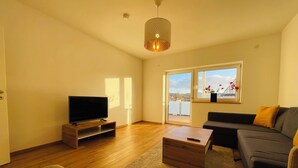 Un soggiorno moderno con una TV a schermo piatto su un mobile in legno, un divano grigio, un tavolino in legno e un'ampia finestra con vista su un balcone.