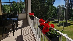 A covered outdoor area with a table and chairs, a railing with potted plants, and a view of a green lawn and trees. - Hotel New Bed (Oskarshamn)