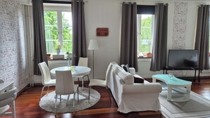 A modern living room with a round dining table, white sofas, a TV on a stand, and large windows with curtains. - Hotel New Bed (Oskarshamn)