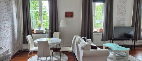 Una sala de estar moderna con una mesa de comedor redonda, sofás blancos, un televisor sobre un mueble bajo y ventanas amplias con cortinas.