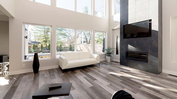 A spacious living room with a white sofa, a flat-screen TV mounted on a dark wall, and a fireplace. Large windows provide natural light and views of the outdoors.