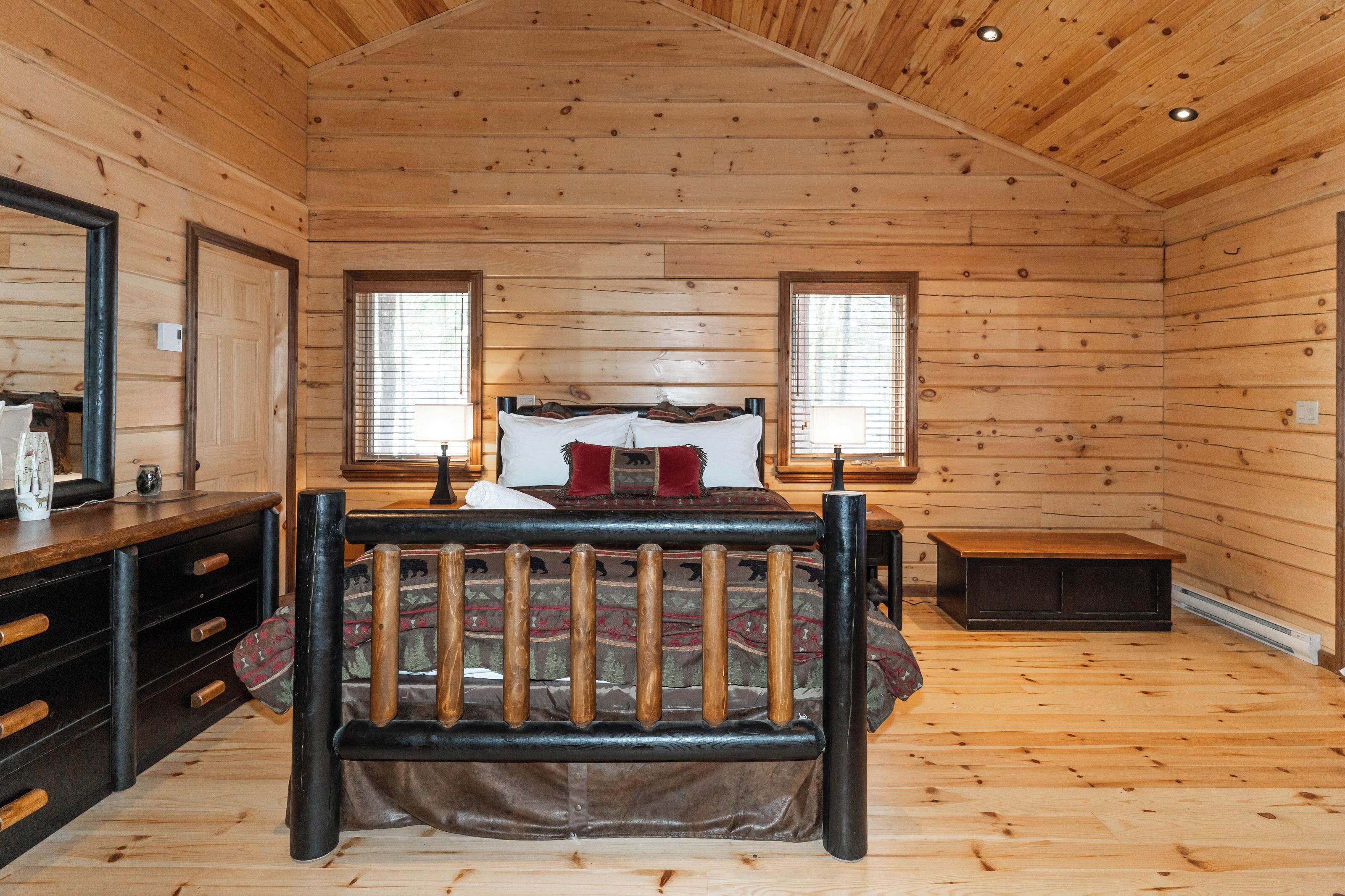 A wooden cabin bedroom with a bed, dresser, and two windows.