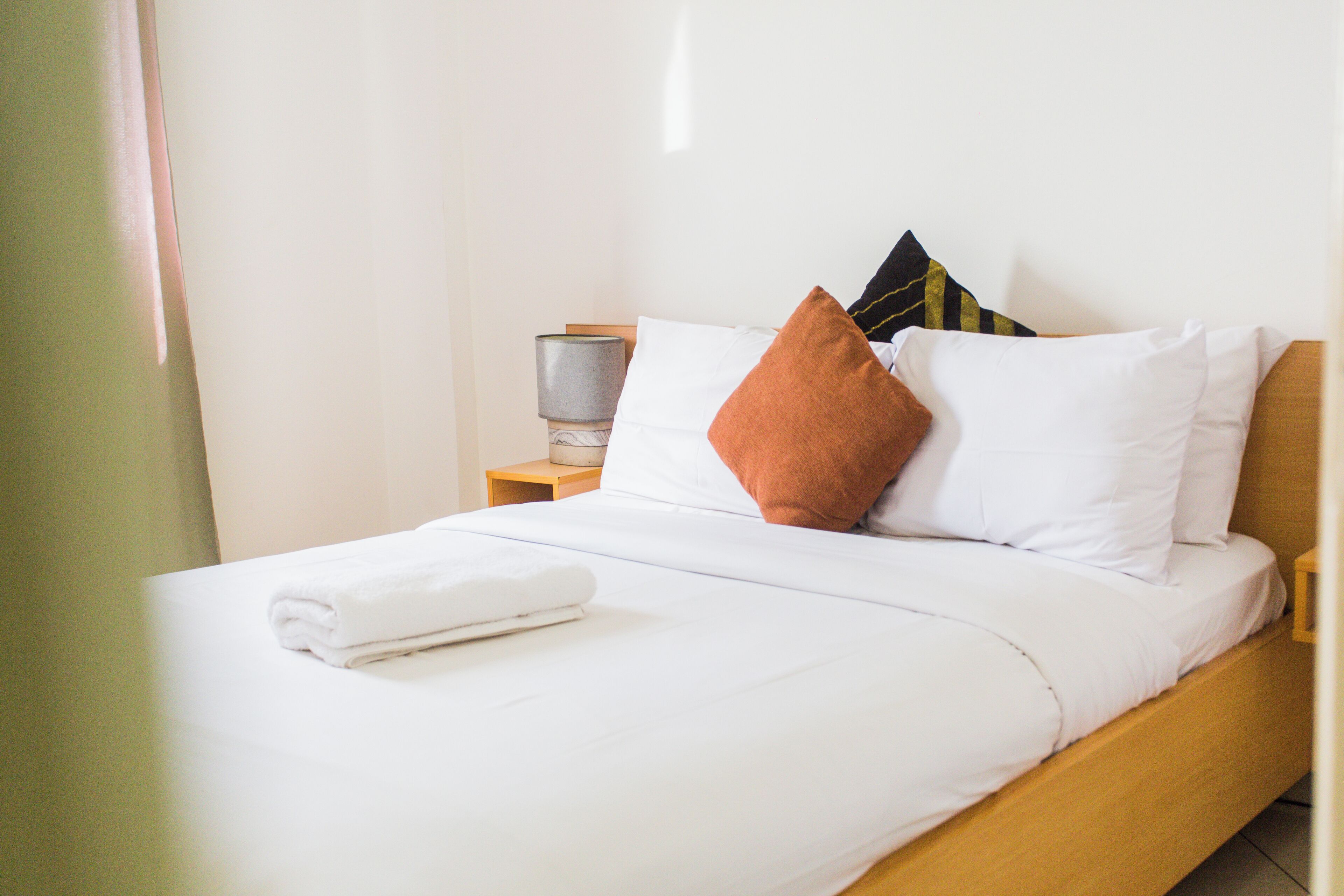 A neatly made bed with white linens and a folded towel, a wooden nightstand with a lamp, and a television on the nightstand.