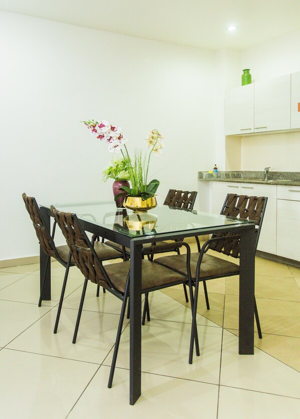A dining area with a glass table surrounded by six chairs, a vase with flowers, and a kitchen in the background. - Accra Luxury Apartments at The Clifton (Accra)