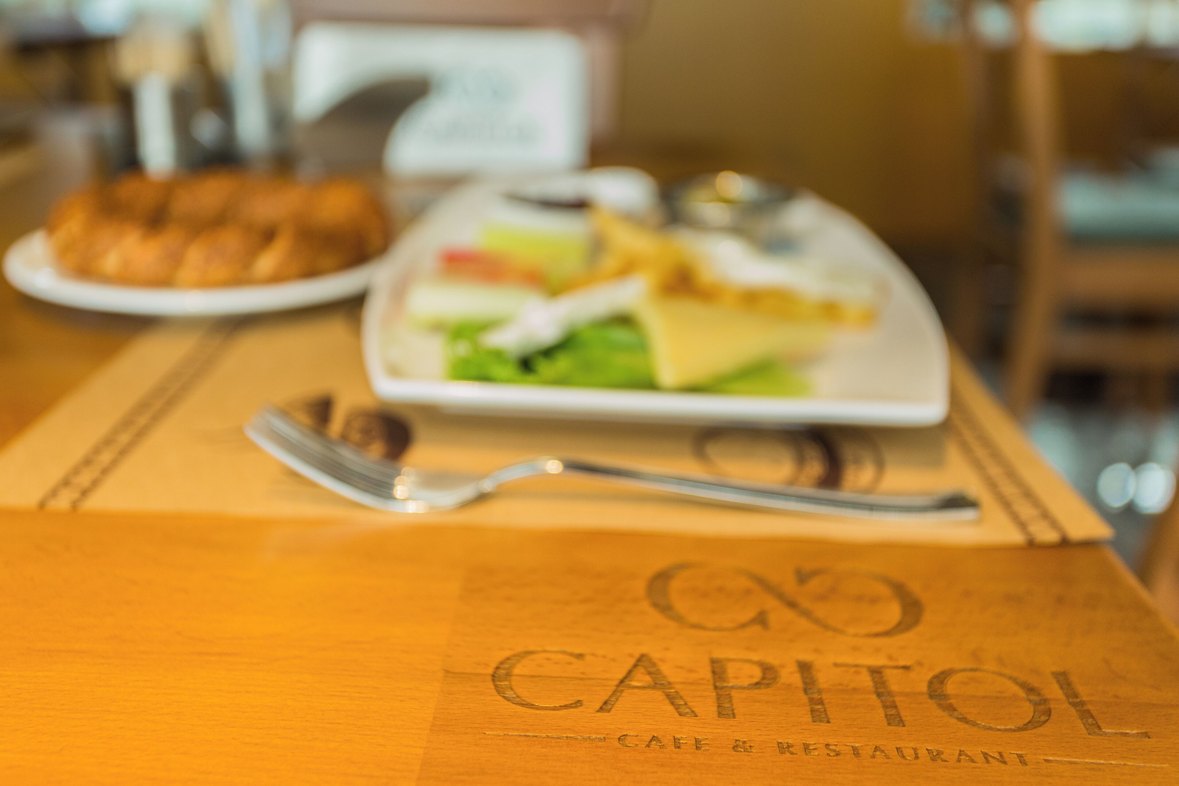 Une table en bois avec une assiette de nourriture et une fourchette. La table porte un logo sur lequel on peut lire « CAPITOL CAFE & RESTAURANT ».