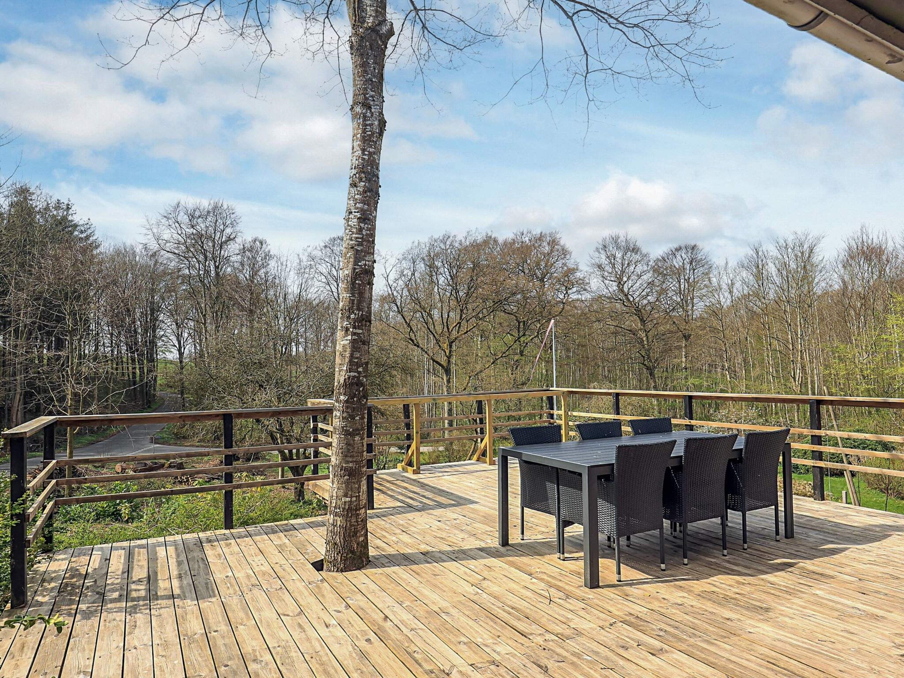 A wooden deck with a table and chairs, surrounded by a railing, overlooking a forested area.