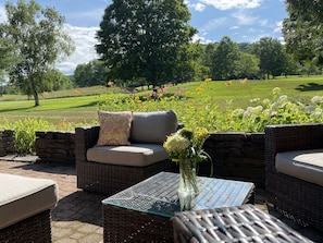 An outdoor seating area with wicker furniture and a glass table, overlooking a well-maintained lawn and trees.