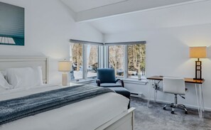 A hotel room with a bed, a desk, a chair, and a window with a view of trees.