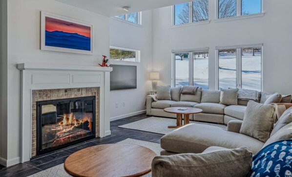 A living room with a fireplace, a large window, and a sofa. - Patio Condo Overlooking Highland Golf, Coach Road (Quechee)