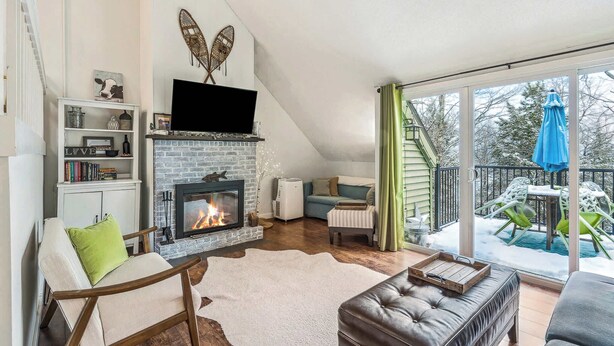 A cozy living room with a fireplace, a TV mounted above it, and a balcony with outdoor seating. - Condo near Ski Hill with Club Access, Kingswood (Quechee)