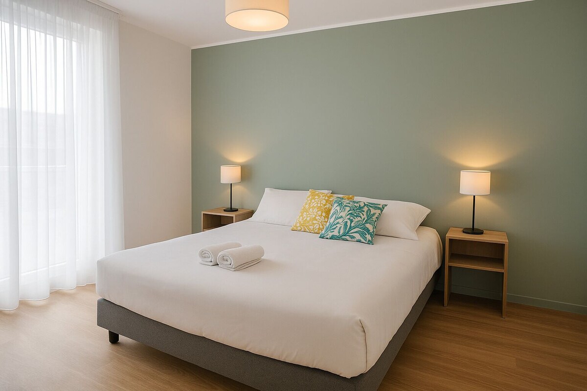 Een net opgemaakt bed met opgerolde handdoeken, twee bedlampen en een raam met dunne gordijnen.