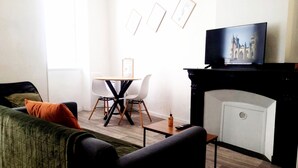 A living room with a sofa, a coffee table, a fireplace, a television, and a dining table with chairs. - Cosy mezzanine studio in Narbonne town center, close to beaches (Narbonne)