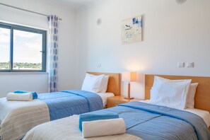 A hotel room with two beds, a window with a view, a painting on the wall, and a nightstand with a lamp. - Lagos Ocean View 3 With Pool by Homing (Lagos)