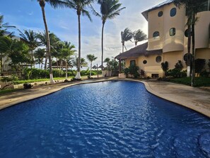 A swimming pool with a curved edge, surrounded by palm trees and a building with multiple windows. - Villas Kaàn (Puerto Morelos)
