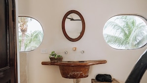 Een landelijke badkamer met een houten wastafel, een spiegel en twee ramen met uitzicht op palmbomen.